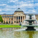 The iconic Kurhaus in Wiesbaden, Germany, captured with a stunning foreground fountain under a bright sky.