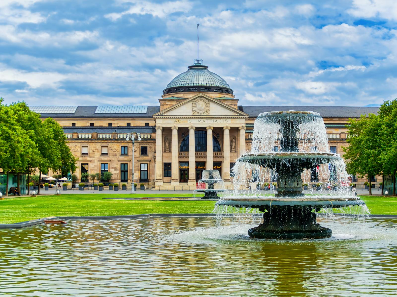 The iconic Kurhaus in Wiesbaden, Germany, captured with a stunning foreground fountain under a bright sky.