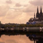 Cologne Cathedral and Hohenzollern Bridge reflecting in the Rhine River under a cloudy sky.