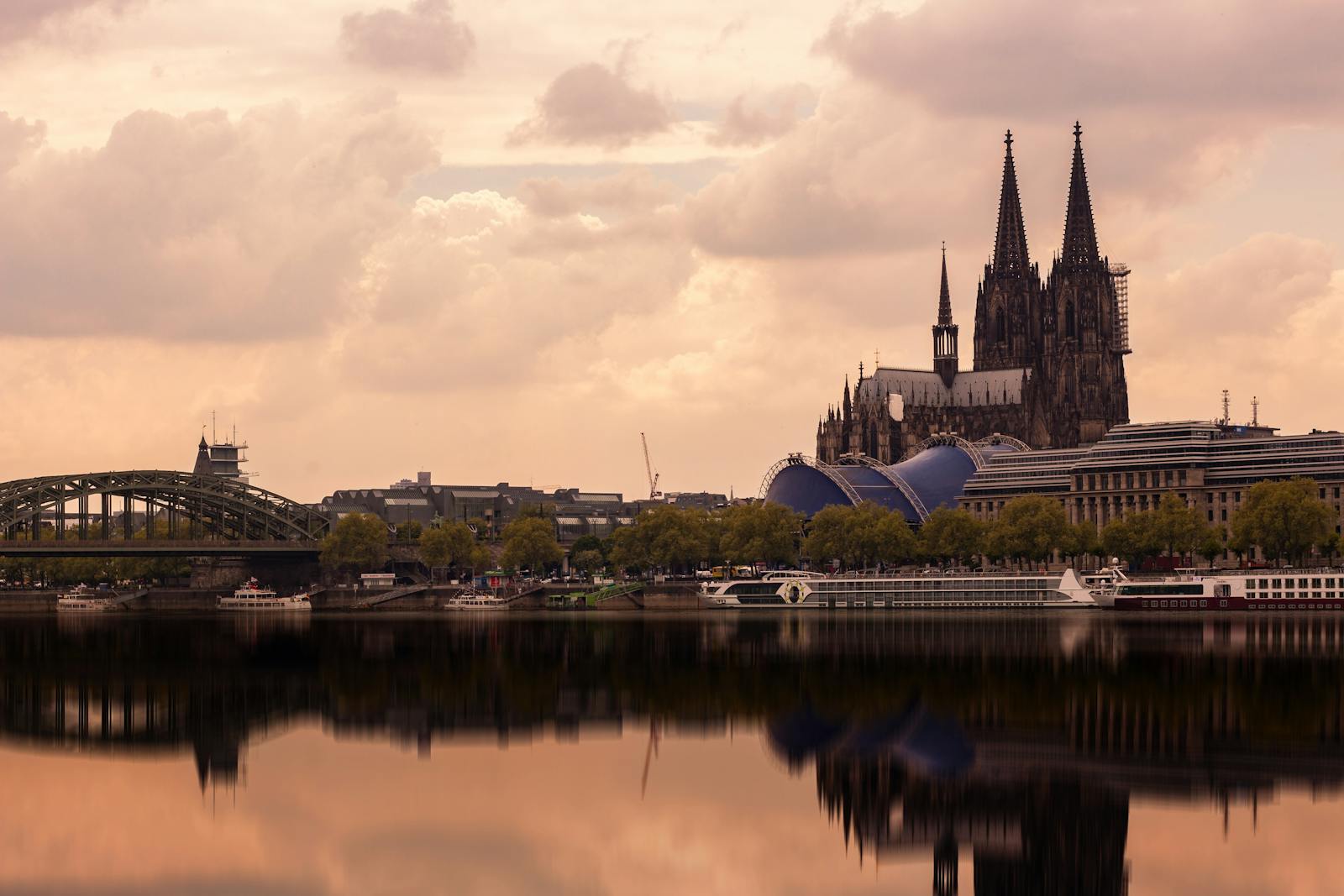 Cologne Cathedral and Hohenzollern Bridge reflecting in the Rhine River under a cloudy sky.